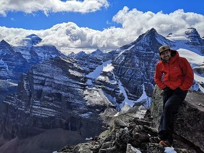 Man in red coat with hat on standing on a summit of beautiful white capped mountains with blue sky and clouds 