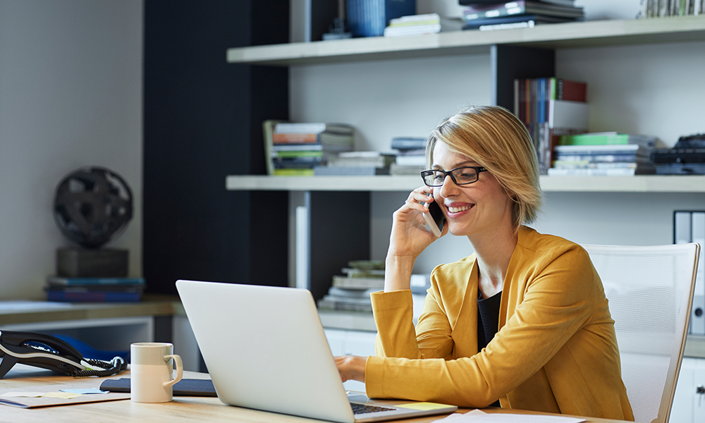 woman in yellow blazer at desk talking on a cell phone and using a computer